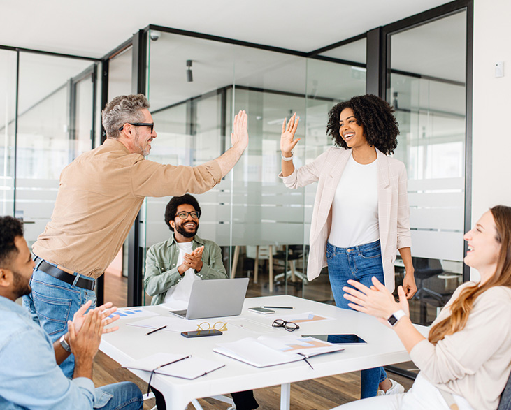 Group of coworkers celebrating success, with two colleagues standing and giving a high five while others applaud around table.