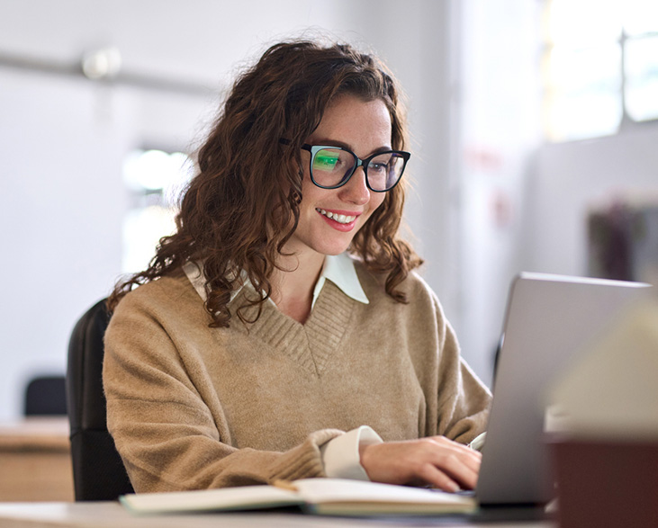 Smiling woman with curly brown hair and glasses working on a laptop