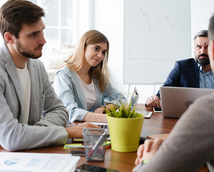 Group of professionals in a serious business meeting around a conference table, with laptops open and a whiteboard showing a line graph