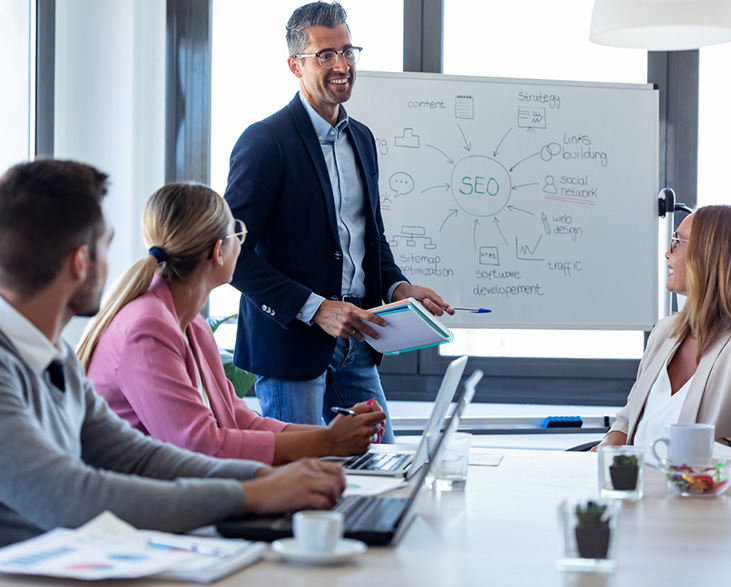 Marketing professional presenting a digital marketing strategy to a team, with an SEO-focused whiteboard diagram and laptops open around the conference table.